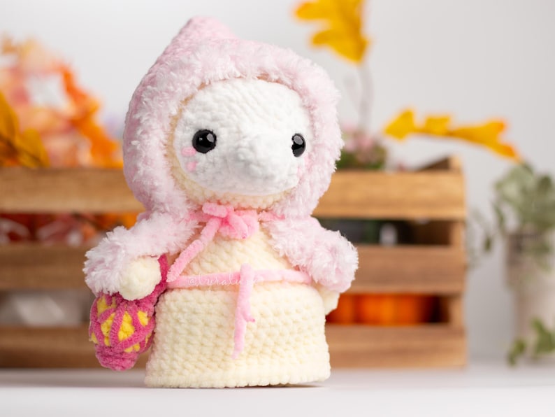 May include: A crocheted plush toy wearing a pink hooded cloak and holding a pink and yellow crocheted basket. The toy has a white body with black eyes and a pink nose.