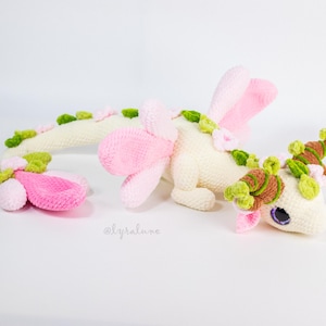 Puede incluir: Tres juguetes de dragón de ganchillo en crema, rosa y verde. El dragón tiene alas rosas, detalles florales verdes y rosas y ojos morados. Los juguetes están hechos a mano y parecen suaves y tiernos.