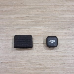 以下が含まれることがあります： 明るい木製の表面に、ドローンまたはカメラ用の2つの黒いアクセサリーが表示されています。1つは長方形のゴムバンドです。もう1つは、DJIのロゴが入った小さな正方形のデバイスです。