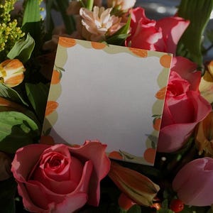 May include: A blank card with a citrus fruit border sits in a bouquet of pink and yellow flowers. The card is white with a green and orange border.
