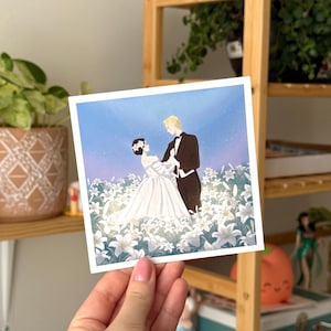 May include: A square art print featuring a couple dancing in a field of white lilies. The bride wears a white gown, and the groom is in a black tuxedo. The background is a light blue sky with a soft glow.