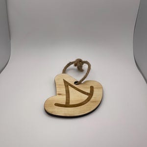 Könnte beinhalten: Herzförmiger Holzschmuck mit einem geschnitzten Design und einer Schlaufe aus Juteschnur zum Aufhängen. Der Schmuck ist aus hellem Holz mit dunklerem Rand gefertigt, und das Design ist ein stilisiertes "D".