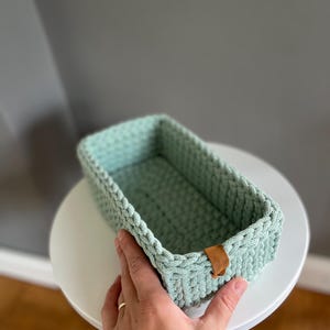 Könnte beinhalten: Ein rechteckiger, hellgrüner Häkelkorb mit geflochtenem Rand und einem kleinen braunen Lederanhänger. Der Korb steht auf einem weißen, runden Tisch. Der Korb ist etwa 25 cm lang, 13 cm breit und 8 cm tief.