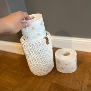 Könnte beinhalten: Ein weißer gehäkelter Toilettenpapierhalter mit zwei Rollen Toilettenpapier mit einem Gänseblümchenmuster.