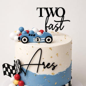 May include: A two-tiered birthday cake with a racing theme. The cake is decorated with blue and white frosting, a checkered flag, and a blue race car. The cake topper reads "TWO fast" in black script. Gold sprinkles and colorful candy balls add to the festive look.
