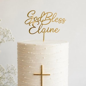 May include: A white cake with a gold cross and a topper that reads "God Bless Elgine." The cake is on a white cake stand and decorated with a textured pattern. The cake is a centerpiece for a religious celebration.