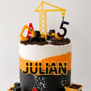 May include: A birthday cake decorated with a construction theme. The cake has layers of white, orange, and black frosting, with the name "JULIAN" in bold letters. Toppings include toy construction vehicles, a crane, a number 5, and a traffic cone.