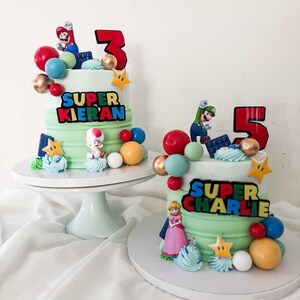 May include: Two birthday cakes with green frosting, decorated with Super Mario characters, colorful spheres, and star-shaped accents. One cake has a red number 3, the other a red number 5, and both have personalized text.