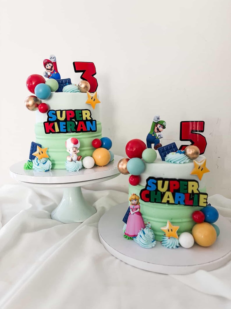 May include: Two birthday cakes with green frosting, decorated with Super Mario characters, colorful spheres, and star-shaped accents. One cake has a red number 3, the other a red number 5, and both have personalized text.