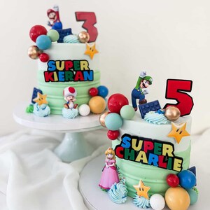 May include: Two tiered cakes with green frosting, decorated with Super Mario characters and colorful candies. One cake has a "3" topper and the text "SUPER KIERAN". The other cake has a "5" topper and the text "SUPER CHARLIE".