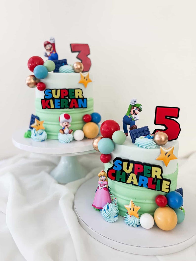 May include: Two tiered cakes with green frosting, decorated with Super Mario characters and colorful candies. One cake has a "3" topper and the text "SUPER KIERAN". The other cake has a "5" topper and the text "SUPER CHARLIE".