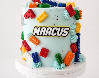 May include: A cylindrical cake with light blue frosting decorated with colorful plastic-style building blocks in red, blue, green, orange, yellow, and white. The name MARCUS is printed in black letters on a yellow background on the front.
