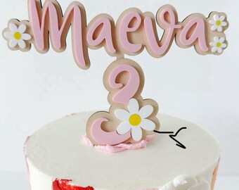 May include: Pink and white wooden cake topper with the name "Maeva" and the number "2" decorated with white daisies.
