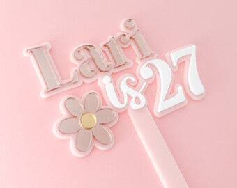 May include: A pink cake topper with the text "Lari is 27" in white and gold lettering. A pink flower with a gold center is below the text.