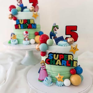 May include: Two birthday cakes with green frosting, decorated with Super Mario characters, colorful spheres, and star-shaped accents. One cake has a red number 3, the other a red number 5. The text "SUPER KIERAN" and "SUPER CHARLIE" are displayed on the cakes.