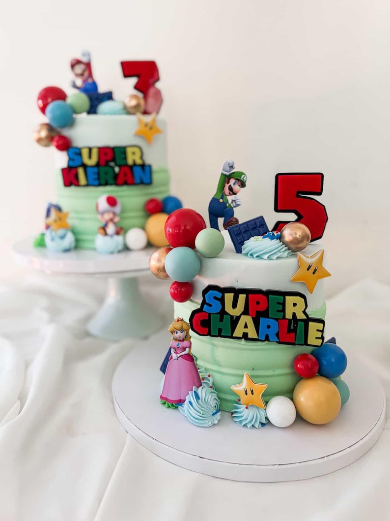 May include: Two birthday cakes with green frosting, decorated with Super Mario characters, colorful spheres, and star-shaped accents. One cake has a red number 3, the other a red number 5. The text "SUPER KIERAN" and "SUPER CHARLIE" are displayed on the cakes.