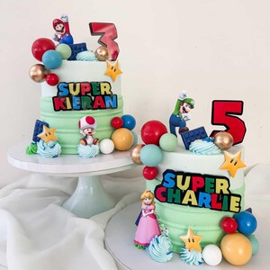 May include: Two birthday cakes with green and white frosting, decorated with Super Mario characters, colorful spheres, and star accents. One cake has a "3" and "Super Kieran" text, the other has a "5" and "Super Charlie" text.