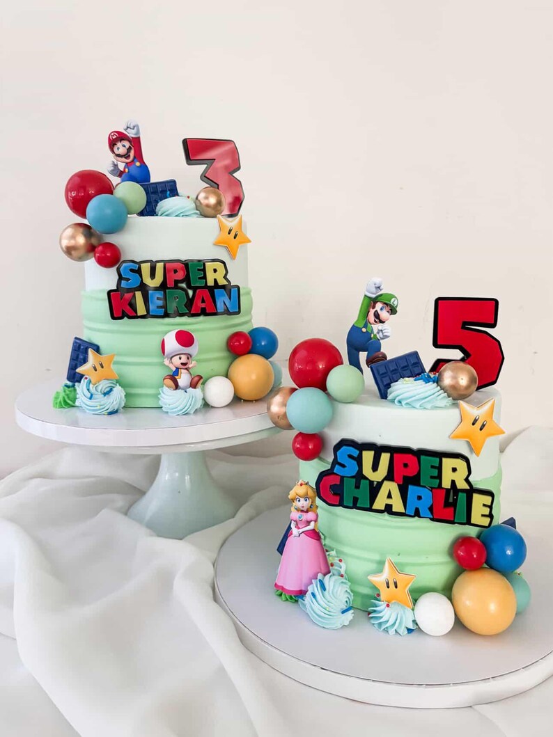 May include: Two birthday cakes with green and white frosting, decorated with Super Mario characters, colorful spheres, and star accents. One cake has a "3" and "Super Kieran" text, the other has a "5" and "Super Charlie" text.