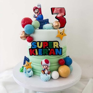 May include: A two-tiered birthday cake with green frosting, decorated with Super Mario characters and colorful spheres. The cake features the text "SUPER KIERAN" and a large number "3". The cake is on a white cake stand.