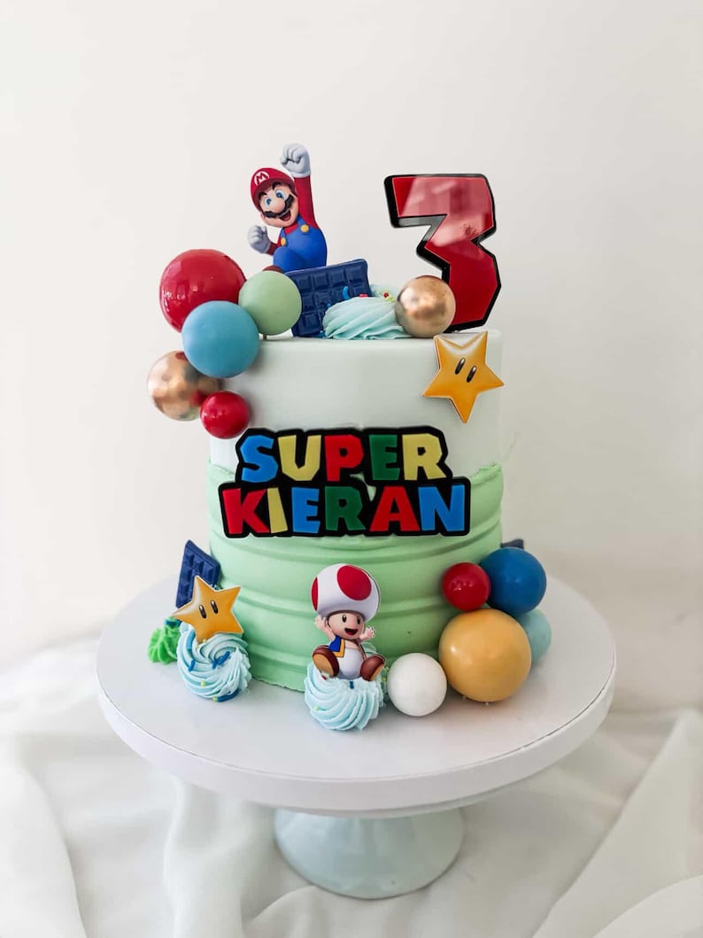 May include: A two-tiered birthday cake with green frosting, decorated with Super Mario characters and colorful spheres. The cake features the text "SUPER KIERAN" and a large number "3". The cake is on a white cake stand.