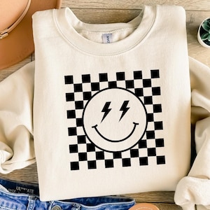 May include: A cream-colored sweatshirt with a black and white checkered pattern and a smiley face with lightning bolts for eyes.