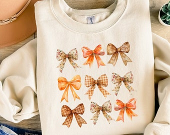 May include: A cream-colored sweatshirt with a pattern of nine orange, brown, and white bows. The bows are arranged in a grid pattern on the front of the sweatshirt.
