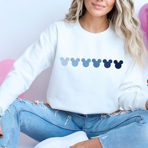 Peut inclure: Un sweat-shirt blanc avec une rangée de cinq têtes de Mickey Mouse bleues imprimées sur le devant. Le sweat-shirt est porté par une personne assise sur une surface blanche avec des ballons en forme de cœur rose et rouge et des pétales de rose.