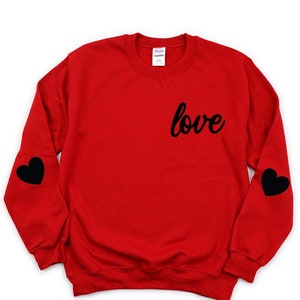 Peut inclure: Sweat-shirt rouge avec des cœurs noirs sur les manches et le mot "love" imprimé sur le devant.