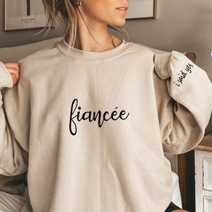 May include: A beige sweatshirt with the text "fiancée" on the front and "i said yes" on the sleeve.
