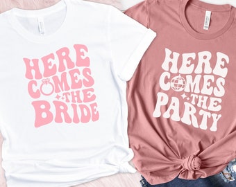 Puede incluir: Dos camisetas blancas con texto rosa. La primera camiseta dice "Here Comes The Bride" con un gráfico de anillo. La segunda camiseta dice "Here Comes The Party" con un gráfico de bola de discoteca.