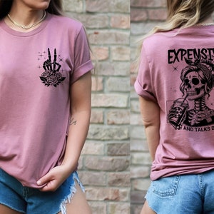 May include: A mauve t-shirt with a black skeleton graphic on the back. The graphic features a skeleton with a bun in her hair, drinking from a cup and the text "Expensive and Talks Back". The front of the shirt has a black graphic of a skeleton hand making a peace sign with roses.