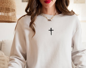 May include: A white sweatshirt with a black cross embroidered on the front. The sweatshirt has long sleeves and a crew neck.