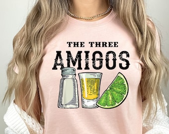 May include: A pink t-shirt with the text "The Three Amigos" in black lettering. The shirt features a graphic of two salt shakers, a shot glass with a yellow liquid, and a lime wedge.