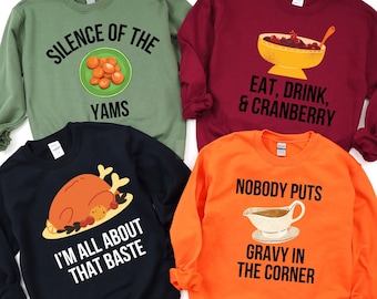 May include: Four different Thanksgiving themed sweatshirts. The first is green with the text "Silence of the Yams" and a plate of yams. The second is maroon with the text "Eat, Drink, & Cranberry" and a bowl of cranberry sauce. The third is navy blue with the text "I'm all about that baste" and a roasted turkey. The fourth is orange with the text "Nobody puts gravy in the corner" and a gravy boat.