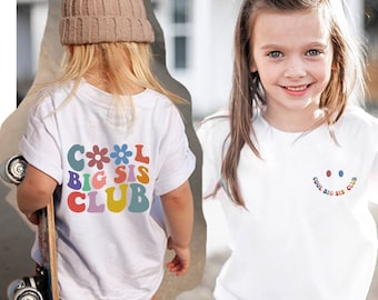 Może przedstawiać: Dwoje dzieci w białych koszulkach z napisem "Cool Big Sis Club" w kolorowych literach. Jedno dziecko ma na sobie brązowy kapelusz i trzyma deskorolkę. Drugie dziecko się uśmiecha.