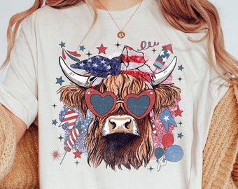 Könnte beinhalten: Ein weißes T-Shirt mit einem Aufdruck einer Highland-Kuh, die eine rote Herz-Sonnenbrille und ein Bandana mit der amerikanischen Flagge trägt. Die Kuh ist von Sternen, Feuerwerk und Ballons umgeben.