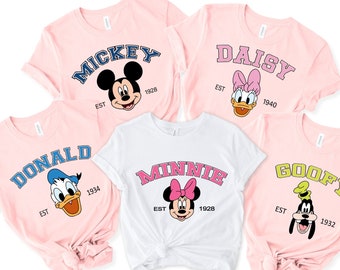 Könnte beinhalten: Fünf rosa und weiße T-Shirts mit verschiedenen Disney-Charakteren bedruckt. Die Charaktere sind Mickey Mouse, Minnie Mouse, Donald Duck, Daisy Duck und Goofy. Jedes Shirt trägt den Namen des Charakters und das Jahr seiner Gründung.