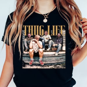 May include: Black t-shirt with a graphic print of four people sitting on a porch. The text "THUG LIFE" is printed in gold letters above the image.