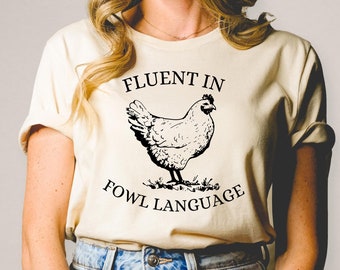 Może przedstawiać: Beżowa koszulka z czarnym nadrukiem kury i tekstem "Fluent in Fowl Language".