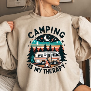 Op de afbeelding: Een beige sweatshirt met een retro-stijl grafische afbeelding van een camper in een bos met de tekst "Camping is my therapy".