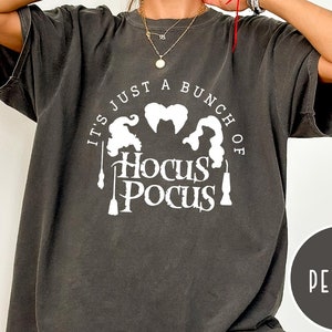 May include: A dark gray t-shirt with a white graphic print of the Sanderson sisters from the movie Hocus Pocus. The text on the shirt reads "It's just a bunch of Hocus Pocus".