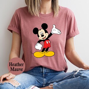 Puede incluir: Camiseta color malva con un dise&ntilde;o de Mickey Mouse. El personaje de dibujos animados se muestra con orejas negras, una mano enguantada blanca, pantalones cortos rojos, zapatos amarillos y una cara sonriente. Prenda de vestir informal.
