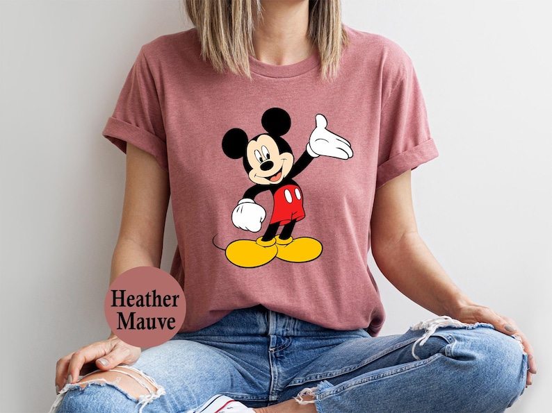 Puede incluir: Camiseta color malva con un dise&ntilde;o de Mickey Mouse. El personaje de dibujos animados se muestra con orejas negras, una mano enguantada blanca, pantalones cortos rojos, zapatos amarillos y una cara sonriente. Prenda de vestir informal.