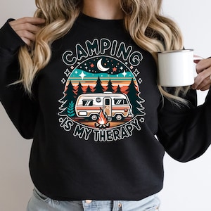 Op de afbeelding: Zwart sweatshirt met een retro-grafische afbeelding van een camper in een bos met de tekst "Camping is my therapy".