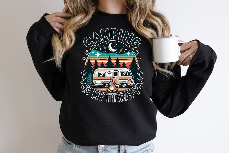 Op de afbeelding: Zwart sweatshirt met een retro-grafische afbeelding van een camper in een bos met de tekst "Camping is my therapy".