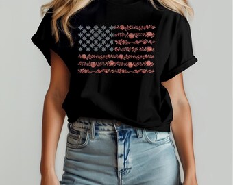 Könnte beinhalten: Schwarzes T-Shirt mit einem floralen Design der amerikanischen Flagge. Die Sterne der Flagge sind hellblau, und die Streifen bestehen aus roten und rosa Blumen und Blättern. Das T-Shirt hat einen Rundhalsausschnitt.