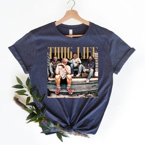 May include: Navy blue t-shirt with a graphic of four people sitting on a porch. The graphic includes the text "THUG LIFE" in gold letters.