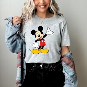 Puede incluir: Una camiseta gris claro con un gr&aacute;fico colorido de Mickey Mouse. Mickey se representa con orejas negras, una cara sonriente, guantes blancos, pantalones cortos rojos y zapatos amarillos. La camiseta es de estilo casual.