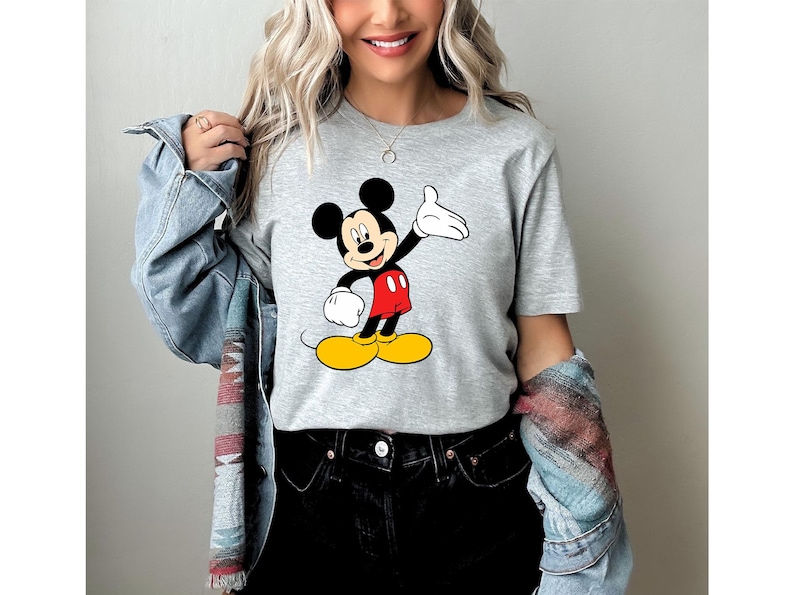 Puede incluir: Una camiseta gris claro con un gr&aacute;fico colorido de Mickey Mouse. Mickey se representa con orejas negras, una cara sonriente, guantes blancos, pantalones cortos rojos y zapatos amarillos. La camiseta es de estilo casual.