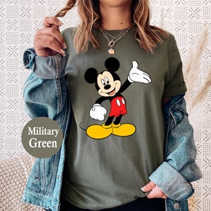 Puede incluir: Camiseta verde militar con un dise&ntilde;o de Mickey Mouse. Mickey se muestra con orejas negras, guantes blancos, pantalones cortos rojos y zapatos amarillos. El conjunto se completa con una chaqueta vaquera. Tambi&eacute;n es visible el texto "Military Green".